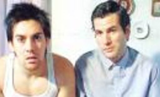 Dos hombres sentados, mirando al frente con cara de sorpresa, se encuentran dentro de una casa. La luz es amplia.