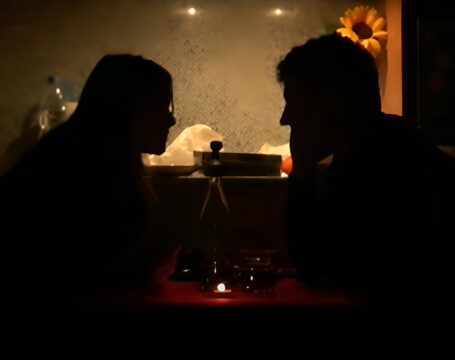 Imagen de pareja teniendo una cena romántica a la luz de las velas en el salón/cocina de su casa. Al fondo se ve la campana extractora de la cocina y un girasol que decora la imagen. La diferencia de brillo entre el fondo y cena provoca que solo se vea la silueta de la pareja y la mesa.