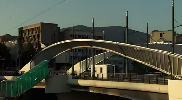 Una vista de un moderno puente peatonal arqueado, con una pasarela verde que desciende hacia el lado izquierdo, bajo un cielo despejado. Edificios y montañas se ven al fondo.