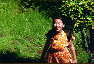 Una chica en primer plano con una selva en plano general ella lleva un vestido naranja con manchas blancas y esta sonriendo con los ojos cerrados