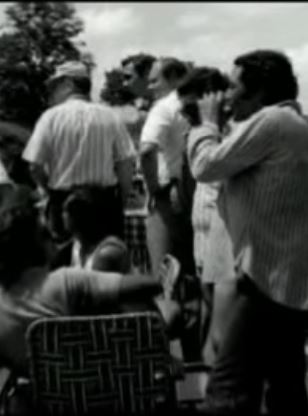 Grupo de personas observando y grabando con una cámara en un espacio exterior durante un evento o reunión.