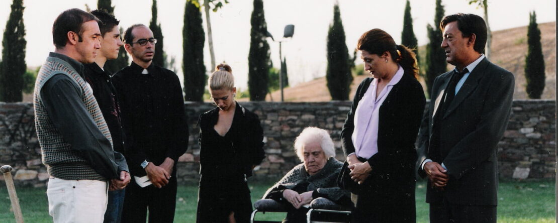 Fotograma de El último adiós. Grupo de personas en medio círculo con la cabeza gacha vestidos de negro. IEntre ellos familiares, amigos y un cura en el exterior de un funeral.