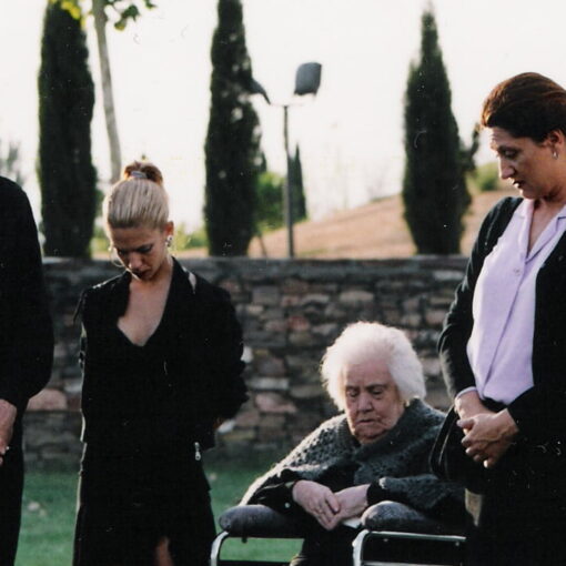 Fotograma de El último adiós. Grupo de personas en medio círculo con la cabeza gacha vestidos de negro. IEntre ellos familiares, amigos y un cura en el exterior de un funeral.