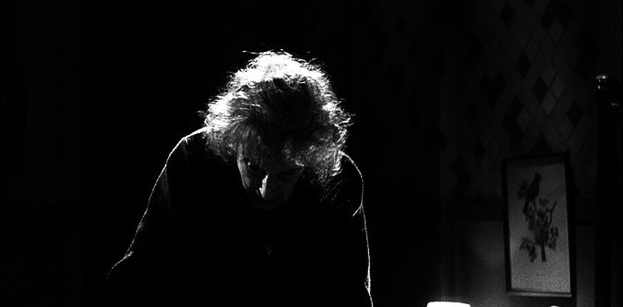 Imagen en blanco y negro de una persona mayor, encorvada y parcialmente a contraluz, en una habitación oscura con una lámpara encendida y un cuadro en la pared.