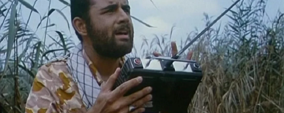 Imagen de uno de los protagonistas en el campo, rodeado de vegatación. Viste un fular de cuadros y una camiseta de manga corta tiene el pelo corto rizo y una barba prominente. está mirando al cielo y en sus manos tienen un controlador negro con una antena metálica que emplea para pilotar el dron.