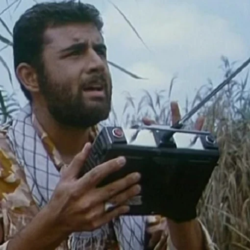 Imagen de uno de los protagonistas en el campo, rodeado de vegatación. Viste un fular de cuadros y una camiseta de manga corta tiene el pelo corto rizo y una barba prominente. está mirando al cielo y en sus manos tienen un controlador negro con una antena metálica que emplea para pilotar el dron.