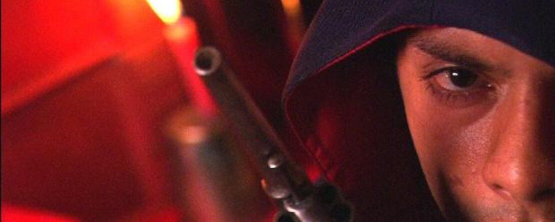 Primer plano de una persona, a la cual solo se le puede ver la mitad de la cara. Lleva una capucha puesta y al lado tiene una pistola. La luz es débil, ya que es una iluminación artificial.