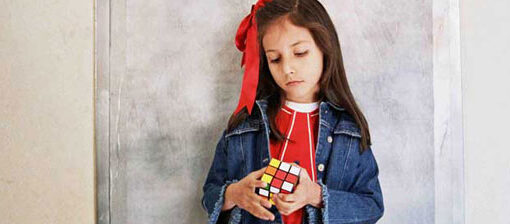 Fotograma de No me gustan los peces de colores. La imagen muestra a una niña de pie frente a una pared gris. Lleva un vestido rojo y una chaqueta azul. En sus manos sostiene un cubo de Rubik.