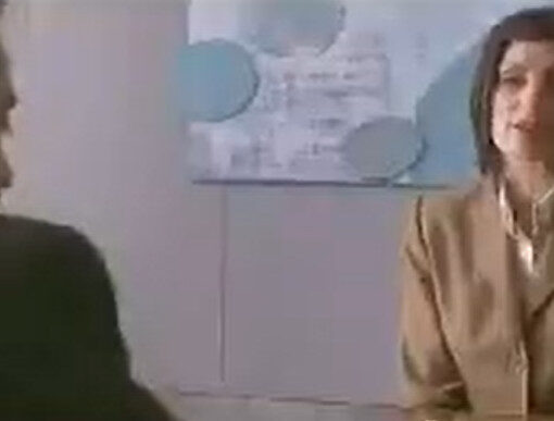 > Una mujer habla con un hombre. Ella lleva un traje beige y mira de frente; el hombre da la espalda.