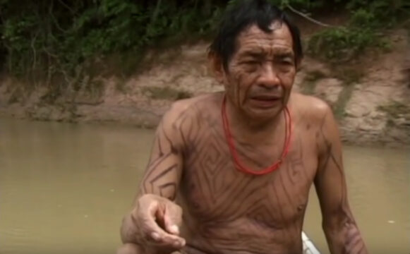 Xina Bena Nuevos tiempos. En el fotograma del documental se ve a un hombre indígena con el torso tatuado, sentado en una canoa sobre un río, rodeado de selva.