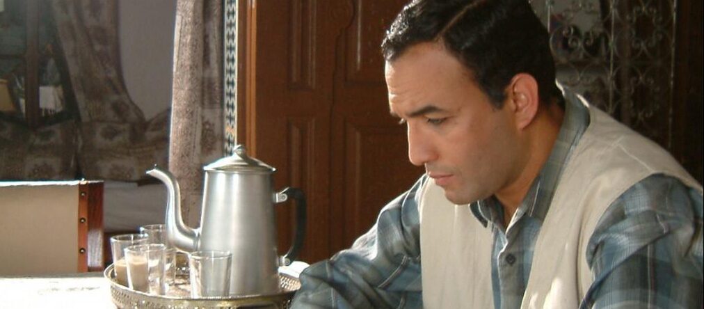 Hombre de mediana edad, en una habitación. Con aspecto pensativo sentado en una mesa al lado de una tetera metálica.
