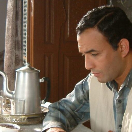 Hombre de mediana edad, en una habitación. Con aspecto pensativo sentado en una mesa al lado de una tetera metálica.