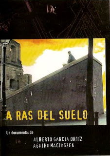 En el centro, lo que parece una rampa de cemento y en lo alto de esa una persona sentada. en la parte izquierda un edificio en ruinas. Sobre esto el titulo A RAS DEL SUELO en color amarillo y ubicado en la parte inferior.