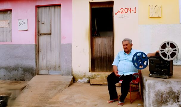 Un hombre de mediana edad se sienta en una silla junto a un proyector de cine antiguo al aire libre. La escena transcurre frente a dos casas con puertas de madera y paredes de colores pastel.