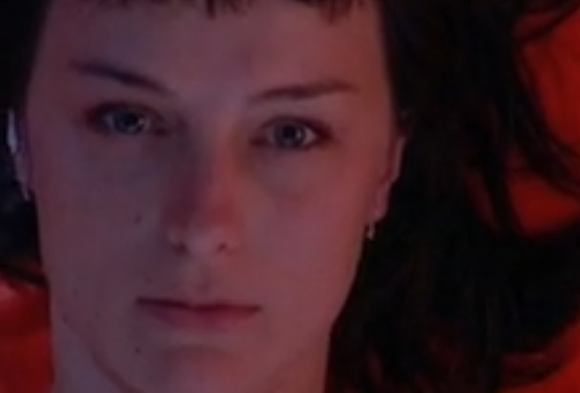 Rostro inexpresivo de una mujer tumbada sobre un colchón rojo.