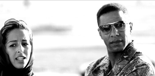 Fotografía en blanco y negro de una mujer con velo hablando, junto a un hombre con gafas oscuras y camisa estampada, que la observa con los brazos cruzados.