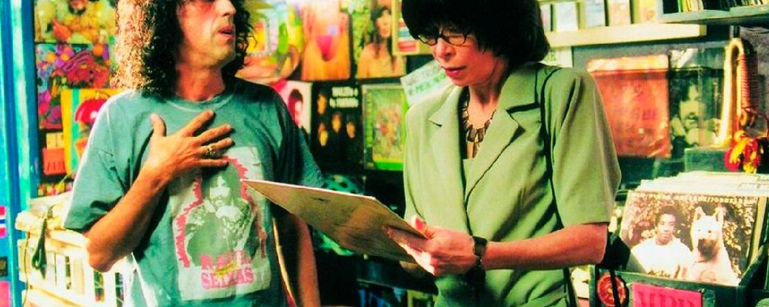 Un hombre y una mujer conversan en una tienda de discos, rodeados de portadas coloridas y vinilos.