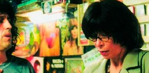 Un hombre y una mujer conversan en una tienda de discos, rodeados de portadas coloridas y vinilos.