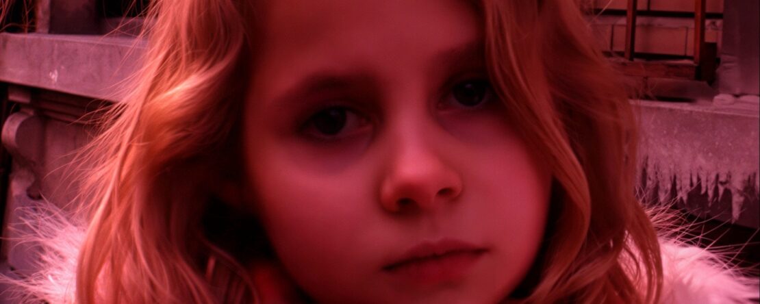 Primer plano de una niña rubia que está mirando directamente a cámara, tiene una expresión triste. La imagen tiene un tono rojizo