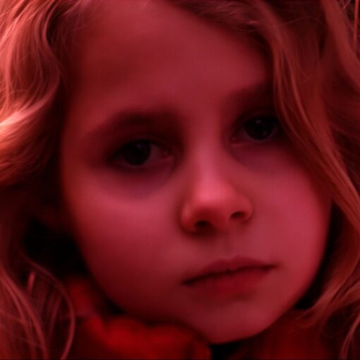 Primer plano de una niña rubia que está mirando directamente a cámara, tiene una expresión triste. La imagen tiene un tono rojizo