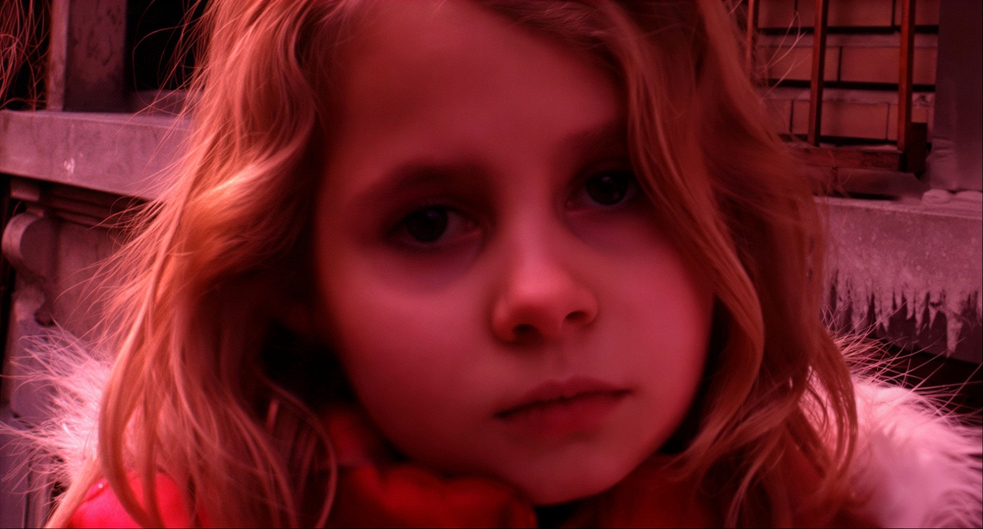 Primer plano de una niña rubia que está mirando directamente a cámara, tiene una expresión triste. La imagen tiene un tono rojizo