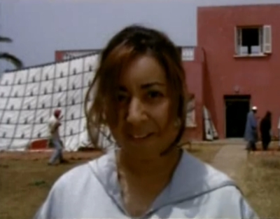 Mujer vestida de blanco mirando fijamente hacia el espectador, con una zona semi urbana marroquí de tonos rojizos de fondo.