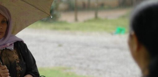 Dos mujeres conversan al aire libre. Una de ellas con hijab, al frente y de espaldas, mira a la otra, que sostiene un paraguas y lleva un pañuelo en la cabeza. El ambiente es tranquilo y parece nublado.
