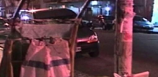 Un hombre recoge cartones en la calle durante la noche, junto a un carrito de reciclaje. A su lado, hay un mueble viejo y desechado cerca de un árbol.