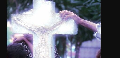 Manos sujetan una cruz translúcida con la figura de Cristo, iluminada Al fondo se ven árboles.