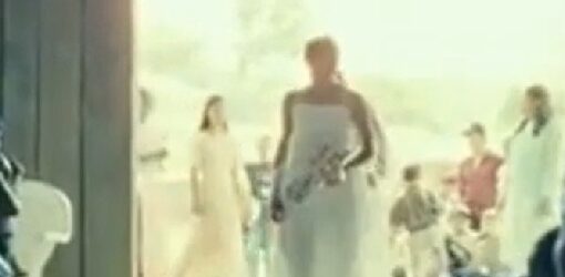 Imagen de una boda; una figura con vestido blanco entra por la puerta mientras los asistentes observan desde los costados en un espacio iluminado por luz natural.
