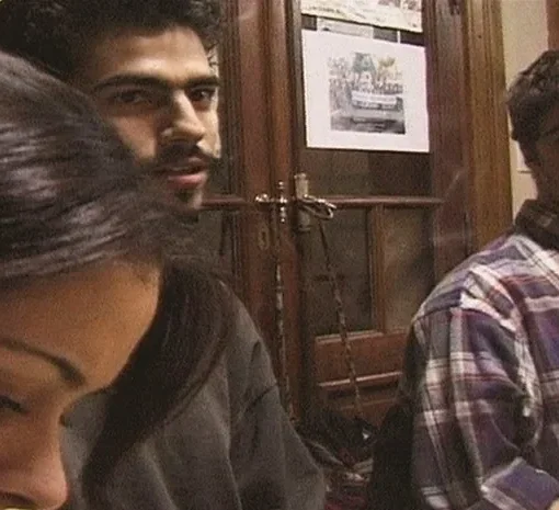 En un interior hay tres personas. A la izquierda, una mujer de cabello largo y oscuro mira hacia abajo. Al centro, un hombre con barba y cabello rizado oscuro mira a la derecha. A la derecha, otro hombre con camisa de cuadros morados mira al centro. Al fondo hay puertas con vidrios, hojas pegadas y cuerdas colgadas.