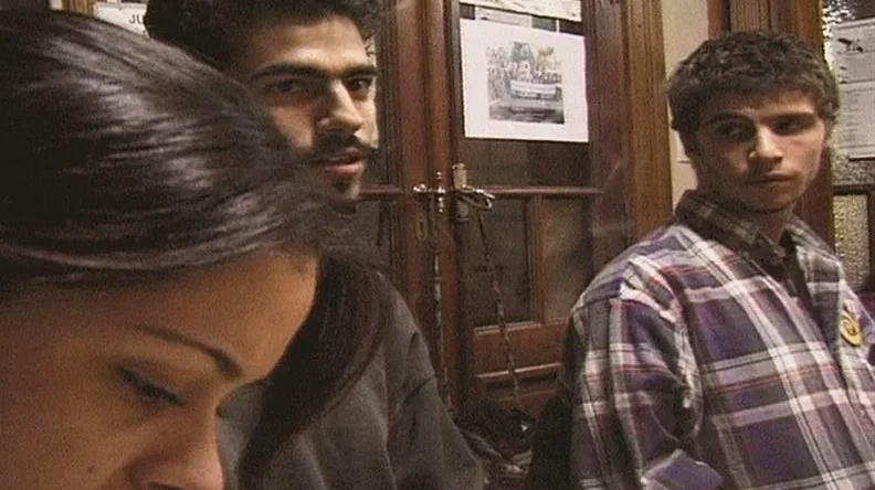 En un interior hay tres personas. A la izquierda, una mujer de cabello largo y oscuro mira hacia abajo. Al centro, un hombre con barba y cabello rizado oscuro mira a la derecha. A la derecha, otro hombre con camisa de cuadros morados mira al centro. Al fondo hay puertas con vidrios, hojas pegadas y cuerdas colgadas.