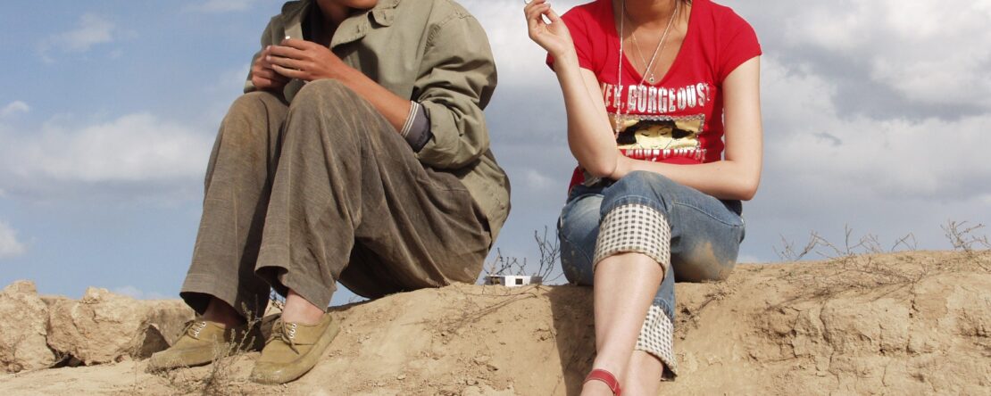 Un chico con ropa tono beige y una chica con una camiseta roja, ambos jóvenes y con rasgos orientales, sentados sobre una roca con el cielo nuboso de fondo.