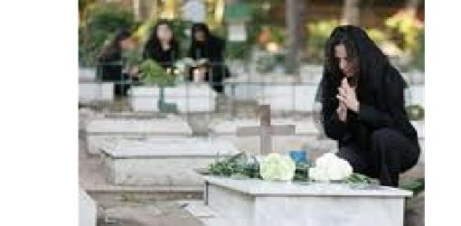 Mujer vestida de negro reza de rodillas junto a una tumba con flores en un cementerio; al fondo, otras personas también de luto.