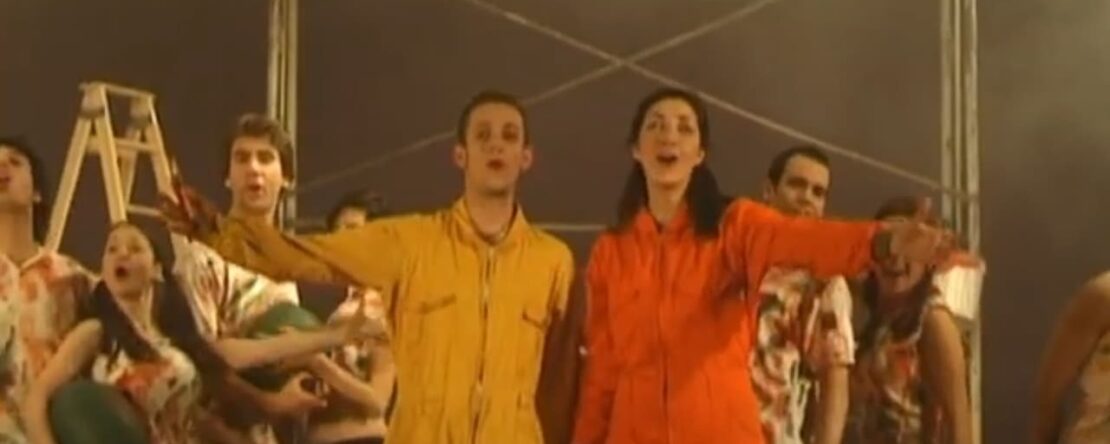 En primer plano, un hombre vestido de amarillo y una mujer vestida de naranja. Ambos están cantando y detrás hay un grupo de personas que les acompañan. La luz es neutral.