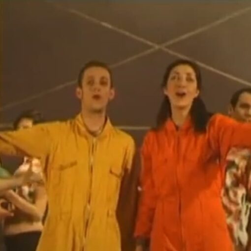 En primer plano, un hombre vestido de amarillo y una mujer vestida de naranja. Ambos están cantando y detrás hay un grupo de personas que les acompañan. La luz es neutral.
