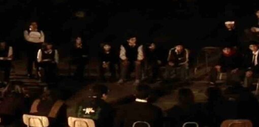 Un grupo de personas, en su mayoría jóvenes, están sentadas en círculo en un espacio oscuro y al aire libre. La escena parece tener un ambiente serio o reflexivo.