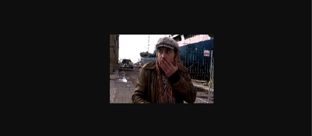 Un hombre con gorra y bufanda colorida se cubre la boca con una expresión de sorpresa en un muelle junto a un barco.