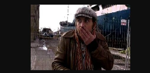 Un hombre con gorra y bufanda colorida se cubre la boca con una expresión de sorpresa en un muelle junto a un barco.
