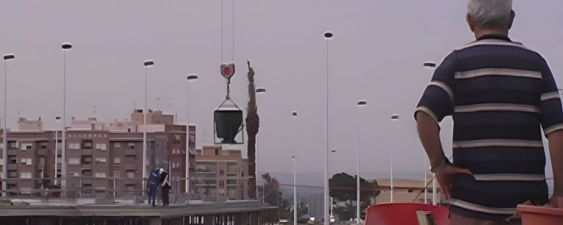 Hombre canoso en la azotea mirando a trabajadores y grúa en un sitio de construcción urbano al atardecer.