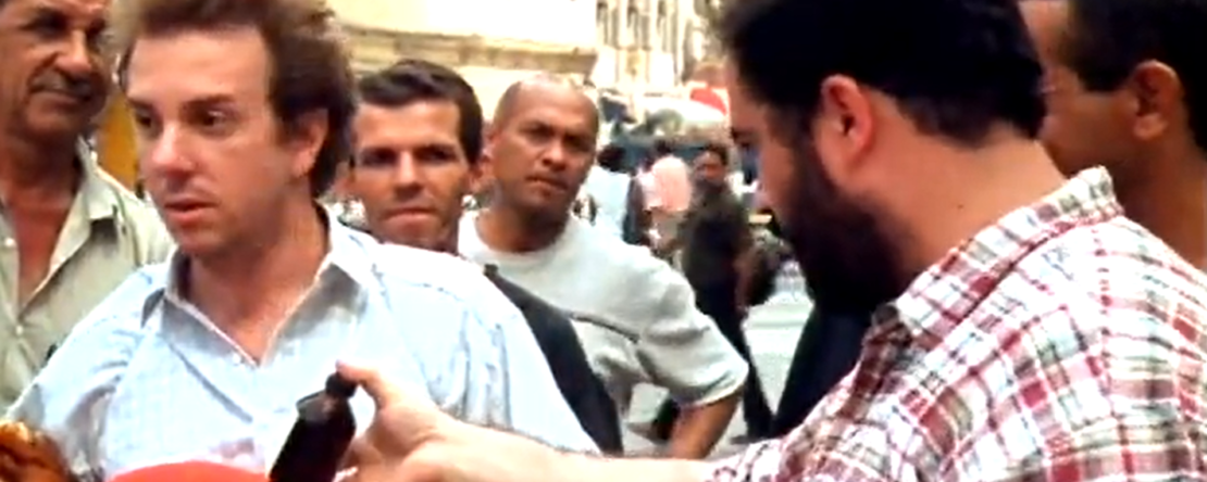 Fotograma de "Trucos, jarabes y otros artículos de confianza". Un vendedor le ofrece jarabe al otro vendedor de peluches en medio de la plaza entre barullo de gente. Miden la confianza.