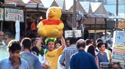 trucos - jarabes y otros Fotograma del cortometraje Trucos, Jarabes y Otros Artículos de Confianza en el que aparece el personaje del vendedor de peluches con las manos alzadas sujetando un gran Winnie de Pooh. Este, trata de venderlo en las calles abarrotadas de la ciudad, a contracorriente entre barullo y gente. Destaca el color amarillo de la camiseta del vendedor y del peluche por encima de la neutralidad del resto del ambiente.