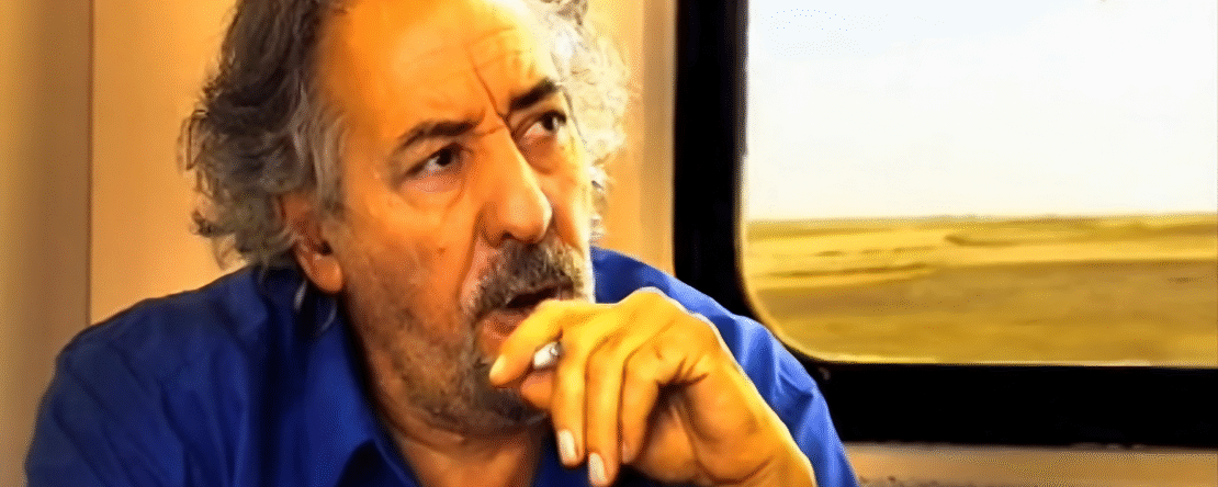 Hombre sentado en un tren, mientras sostiene un cigarro con rostro pensativo