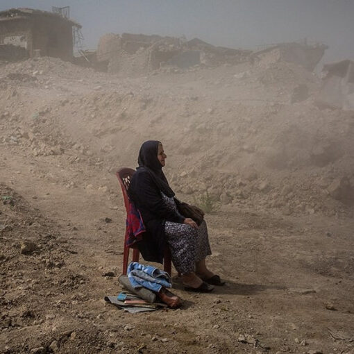 Una mujer sentada en una silla, con una expresión seria, en medio de ruinas y casas destrozadas. El fondo muestra edificios parcialmente destruidos y escombros dispersos por el suelo. La imagen transmite la dureza de la vida en un entorno devastado por el conflicto.