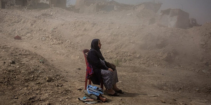Una mujer sentada en una silla, con una expresión seria, en medio de ruinas y casas destrozadas. El fondo muestra edificios parcialmente destruidos y escombros dispersos por el suelo. La imagen transmite la dureza de la vida en un entorno devastado por el conflicto.