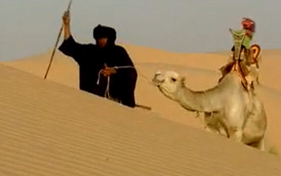 Un hombre con vestiduras negras que se sostiene con una vara caminando a traves del desierto arrastrando un camello con una montura la cual tiene una marioneta encima