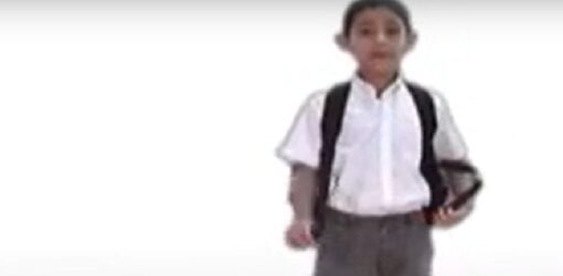 Un niño con uniforme escolar y mochila está de pie sobre un fondo blanco. Sostiene un cuaderno en una mano y mira al frente.