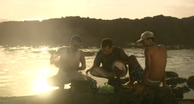 Tres chicos jóvenes en cuclillas en una roca en la orilla del mar, con el sol del atardecer de fondo.