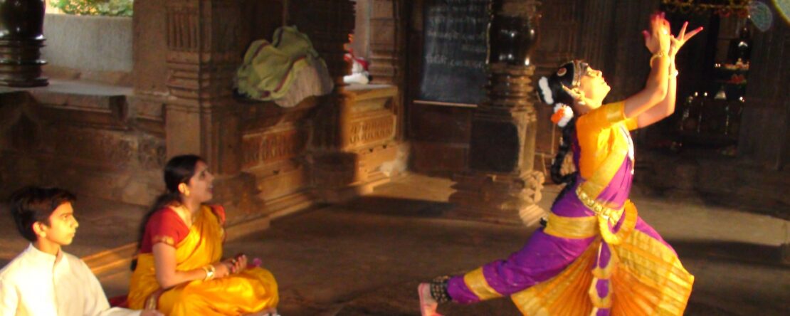 Un niño, con un instrumento, y una niña, asombrada, están sentados mirando a una chica que está bailando. Todos van con ropa llamativa y se encuentran en una especie de templo. La luz del día entra irregularmente por las ventanas.