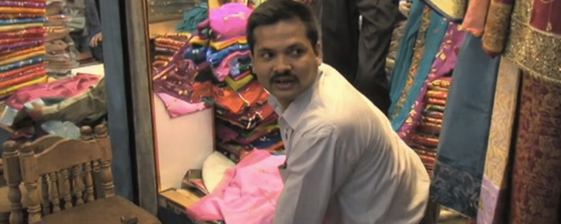 Hombre bangladesí agachado en una tienda de telas, rodeado de vestidos y tejidos de colores, mirando hacia un lado mientras ordena mercancía en un espacio comercial estrecho.